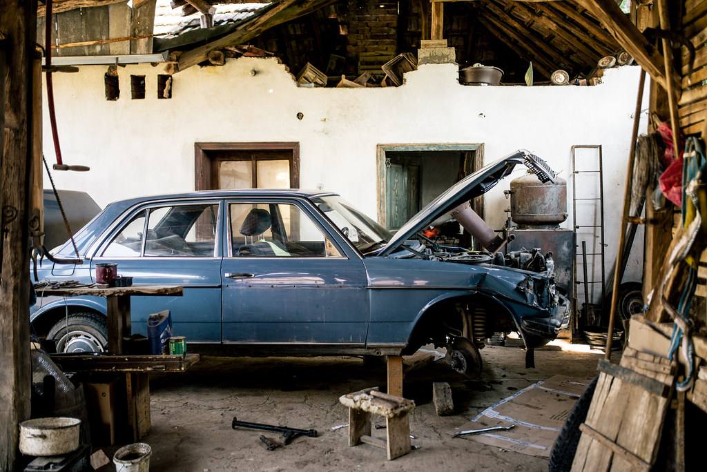Modrý sedan Mercedes-Benz řady 123 v kůlně bez levého předního blatníku, s odkrytým podběhem