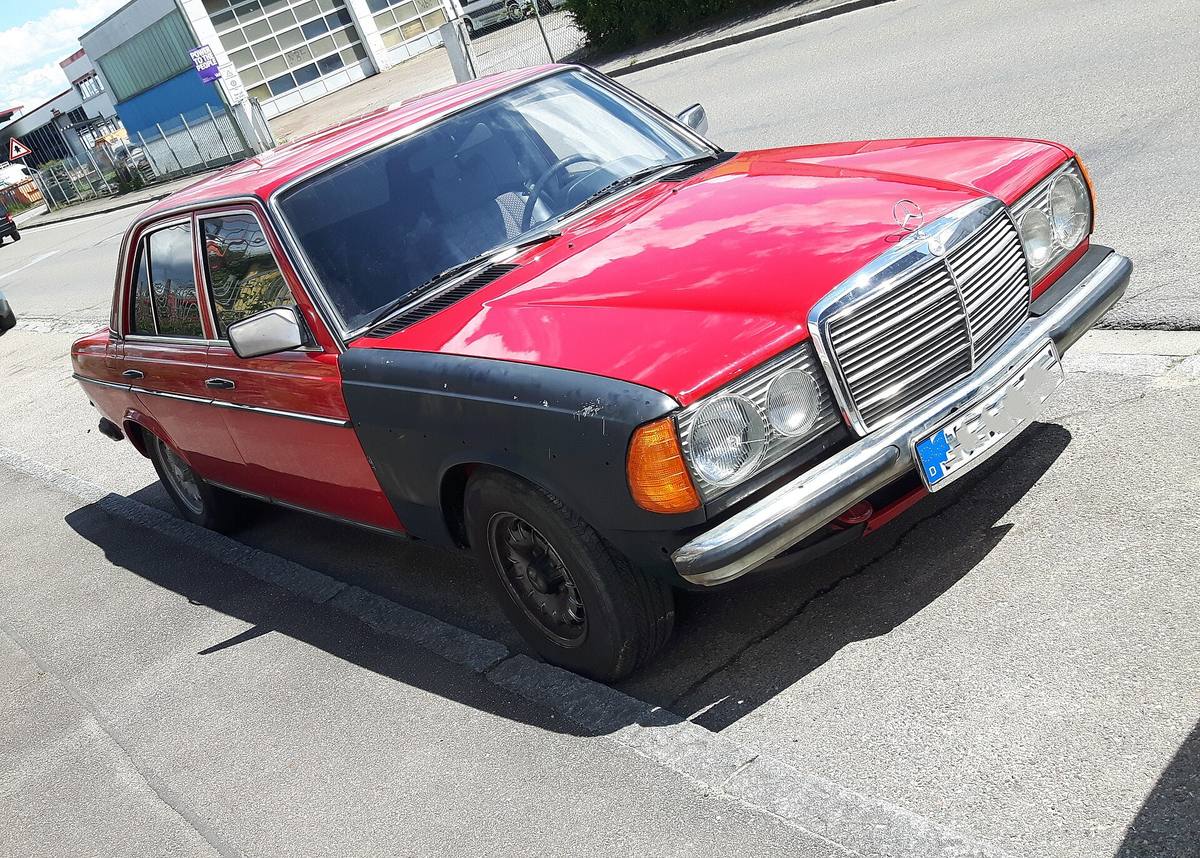 Červený sedan Mercedes-Benz řady 123 s černým nenalakovaným předním blatníkem a černými ocelovými koly stojí u chodníku