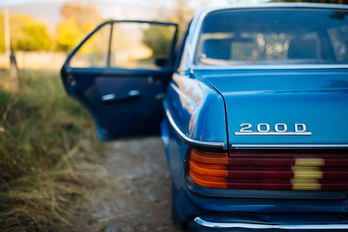 Modrý Mercedes-Benz 200D řady 123 na polní cestě, dveře řidiče pootevřené
